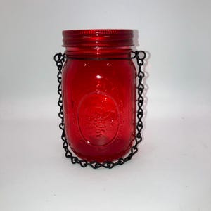 May include: A red glass mason jar with a matching lid and a black chain handle. The jar has an embossed design on the front and is suitable for candles or decorative purposes.