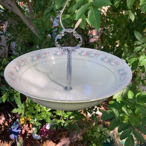 May include: A single-tier serving bowl featuring a cream-colored ceramic base with a delicate floral pattern of pink and blue flowers along the rim. The bowl is attached to a central silver-toned metal handle with a decorative loop top.