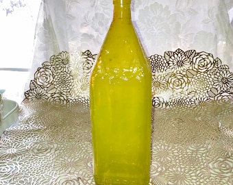 May include: A large, yellow glass bottle with a rounded bottom and a straight neck. The bottle is sitting on a gold-colored, floral-patterned surface.