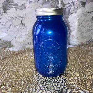 May include: A blue mason jar with a silver lid. The jar has a glossy finish and a decorative embossed design on the front. The jar is sitting on a gold-colored surface.