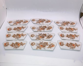 May include: Set of twelve white ceramic tiles with a floral design. Each tile features a brown and orange rose with green leaves. The tiles are shaped like a crescent moon.