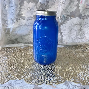 May include: A cobalt blue mason jar with a silver lid. The jar has an embossed design of a crescent moon and stars. The jar is sitting on a gold-colored, floral-patterned placemat.