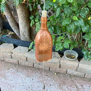 May include: A copper-colored glass bottle wind chime with a woven pattern hanging from a chain. The bottle is suspended from a chain and has a cork stopper.