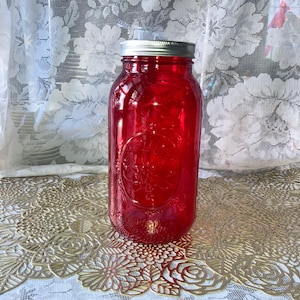 May include: A large, red-tinted glass jar with a silver lid. The jar has a classic mason jar shape with embossed detailing. The jar is sitting on a gold-colored, floral-patterned surface.