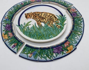 May include: A circular ceramic tile featuring a tiger walking through green grass, surrounded by a ring of twelve separate curved border tiles. Each border tile displays a pattern of green grass, blue accents, and colorful purple, pink, and yellow flowers.