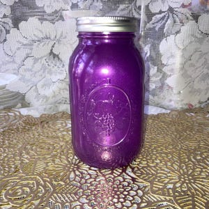 May include: A purple mason jar with a silver lid. The jar has a metallic finish and an embossed design featuring grapes. The jar is sitting on a gold-colored, patterned surface.