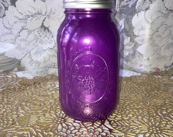 May include: A purple mason jar with a silver lid. The jar has a metallic finish and an embossed design featuring grapes. The jar is sitting on a gold-colored, patterned surface.