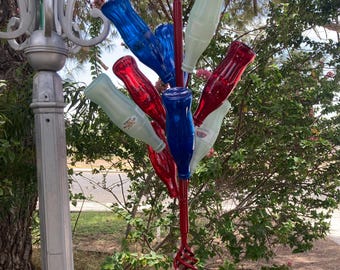 May include: A red metal bottle tree garden stake featuring a central vertical rod with multiple angled branches. Several glass bottles in shades of red, blue, and clear are attached to the branches, creating a colorful display for outdoor decor.