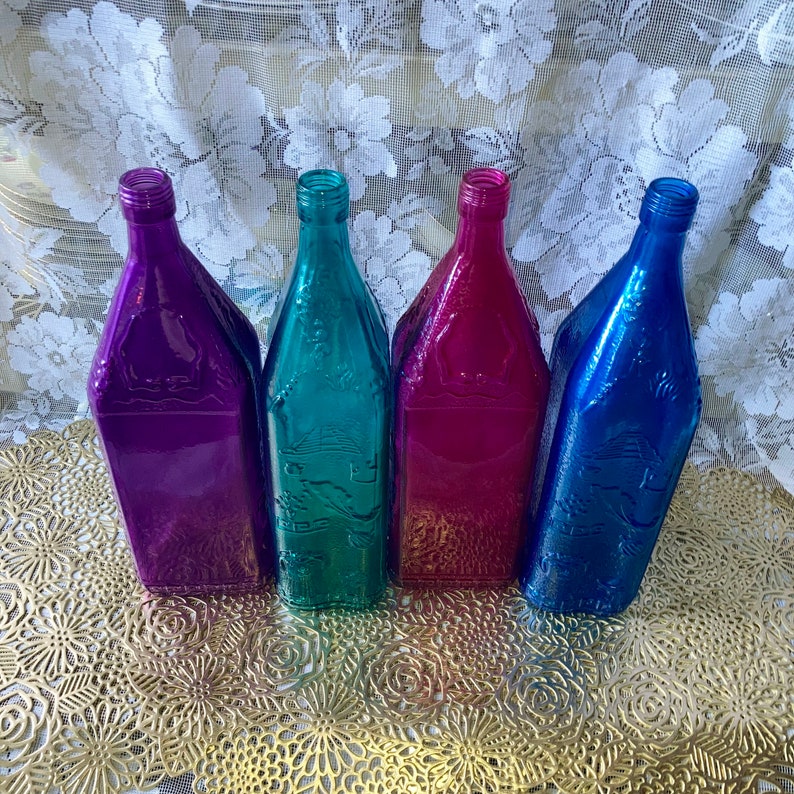 May include: Four glass bottles with a textured surface. The bottles are in shades of purple, teal, and blue. The bottles are sitting on a gold-colored surface.