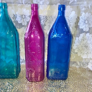 May include: Three glass bottles with a textured surface. The bottles are teal, magenta, and blue.