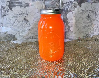 May include: An orange-colored mason jar with a silver lid. The jar has a smooth, glossy finish and a raised design on the front. The jar is sitting on a gold-colored, floral-patterned placemat.