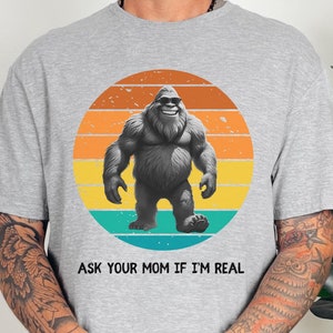 Peut inclure: Un t-shirt gris avec un graphique de coucher de soleil rétro représentant un Bigfoot. Le texte "Ask your mom if I'm real" est imprimé sous le graphique.