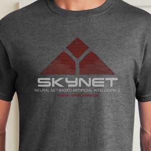 以下が含まれることがあります： 「SKYNET」と「NEURAL NET-BASED ARTIFICIAL INTELLIGENCE」と書かれた赤と白のロゴが入ったダークグレーのTシャツです。