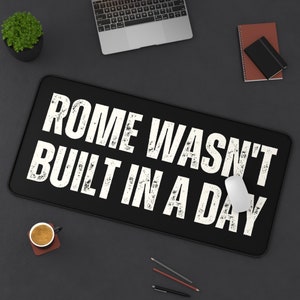 May include: Black desk mat with white text that reads "ROME WASN'T BUILT IN A DAY". A white computer mouse is on the mat.