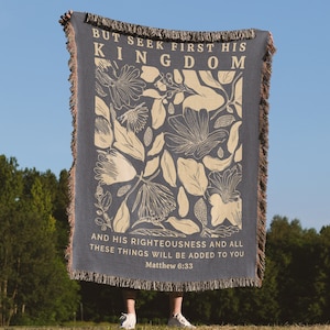 May include: A woven throw blanket with a floral pattern in cream on a dark blue background. The text "BUT SEEK FIRST HIS KINGDOM AND HIS RIGHTEOUSNESS AND ALL THESE THINGS WILL BE ADDED TO YOU Matthew 6:33" is printed in cream on the blanket.