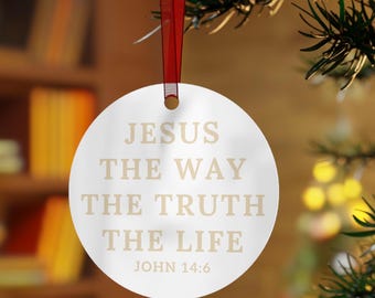 May include: A circular, white Christmas bauble with the inscription "JESUS THE WAY THE TRUTH THE LIFE JOHN 14:6" in a light brown hue. It is suspended by a red ribbon, set against a blurred backdrop of a Christmas tree and a bookcase.