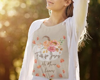 Puede incluir: Camiseta beige con un diseño de calabaza blanca con detalles florales rosas y naranjas. La camiseta tiene el texto "Fall for Jesus He Never Leaves" en un estilo manuscrito, con hojas cayendo.