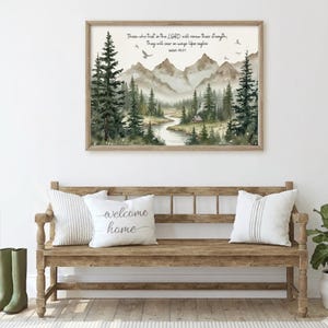 Op de afbeelding: Een ingelijste aquarelprint met een berglandschap met een rivier, bomen en een kleine hut. De tekst "Those who trust in the LORD will renew their strength; They will soar on wings like eagles" staat bovenaan. Een houten bank met kussens staat op de voorgrond.