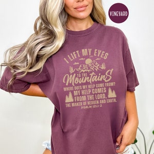 Könnte beinhalten: Ein maroonfarbenes T-Shirt mit einem tannengrünen Grafikdruck von Bergen und Bäumen. Der Text auf dem Shirt lautet "Ich hebe meine Augen zu den Bergen, woher kommt meine Hilfe? Meine Hilfe kommt vom Herrn, dem Schöpfer von Himmel und Erde. Psalm 121:1-2".
