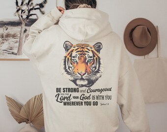 May include: A cream colored hoodie with a tiger graphic on the back. The hoodie has a quote that reads "Be strong and courageous, for the Lord your God is with you wherever you go. Joshua 1:9"