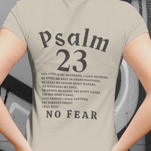 Może przedstawiać: Beżowa koszulka z napisem "Psalm 23" w kolorze czarnym. Tekst kontynuuje werset z Biblii: "Pan jest moim pasterzem, niczego mi nie braknie. Na zielonych pastwiskach daje mi odpoczynek, nad spokojne wody mnie prowadzi. Odnawia moje siły, prowadzi mnie po ścieżkach sprawiedliwości dla swego imienia. Choćbym chodził doliną cienia śmierci, nie ulęknę się zła, bo Ty jesteś ze mną; tw&oacute;j kij i twoja laska są moją pociechą."