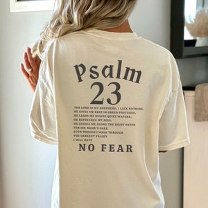 Może przedstawiać: Biały T-shirt z czarnym nadrukiem Psalmu 23. Tekst zawiera werset "Pan jest moim pasterzem, niczego mi nie zabraknie." i "Nie będę się bał."