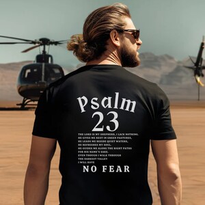 Może przedstawiać: Czarny T-shirt z białym tekstem, kt&oacute;ry brzmi "Psalm 23" i cytatem z wersetu biblijnego. Cytat brzmi: "Pan jest moim pasterzem, niczego mi nie braknie. On sprawia, że odpoczywam na zielonych pastwiskach, prowadzi mnie nad spokojne wody. Przywraca mi siły, prowadzi mnie po właściwych ścieżkach dla swego imienia. Nawet gdybym miał iść przez dolinę cienia śmierci, nie bałbym się zła, bo Ty jesteś ze mną; tw&oacute;j pasterski kij i tw&oacute;j laska mnie pocieszają."