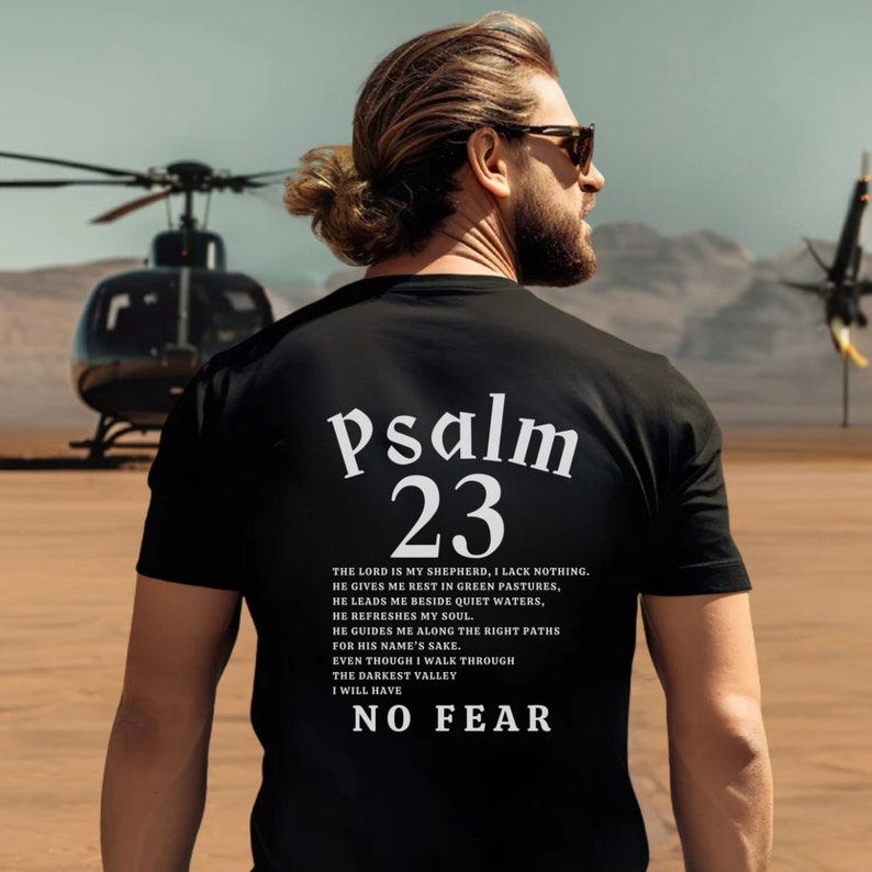 Może przedstawiać: Czarny T-shirt z białym tekstem, kt&oacute;ry brzmi "Psalm 23" i cytatem z wersetu biblijnego. Cytat brzmi: "Pan jest moim pasterzem, niczego mi nie braknie. On sprawia, że odpoczywam na zielonych pastwiskach, prowadzi mnie nad spokojne wody. Przywraca mi siły, prowadzi mnie po właściwych ścieżkach dla swego imienia. Nawet gdybym miał iść przez dolinę cienia śmierci, nie bałbym się zła, bo Ty jesteś ze mną; tw&oacute;j pasterski kij i tw&oacute;j laska mnie pocieszają."