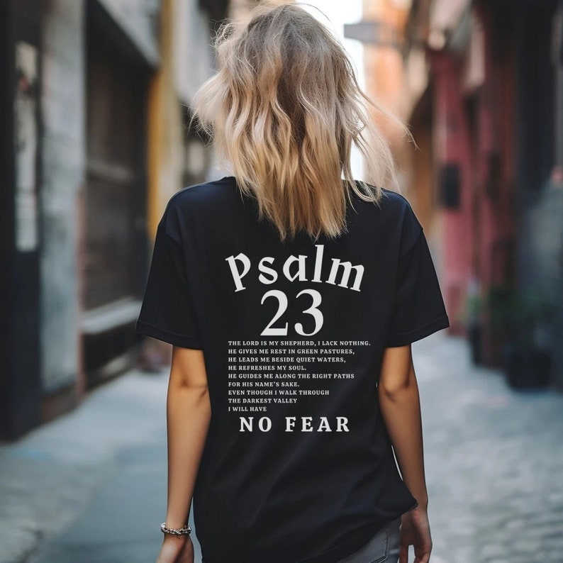 Może przedstawiać: Czarny T-shirt z białym tekstem, kt&oacute;ry brzmi "Psalm 23" i wersetem z Biblii. Werset brzmi: "Pan jest moim pasterzem, niczego mi nie braknie. On sprawia, że odpoczywam na zielonych pastwiskach, prowadzi mnie nad spokojne wody. Odnawia moje siły, prowadzi mnie po prawych ścieżkach dla swego imienia. Nawet gdybym miał iść przez dolinę cienia śmierci, nie bałbym się zła, bo Ty jesteś ze mną; Tw&oacute;j kij i Twoja laska są moim pocieszeniem."
