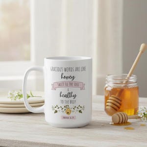 May include: White ceramic mug with the text "Gracious words are like honey, sweet to the soul & healthy to the body" and a floral design. A jar of honey and a honey dipper are next to the mug.