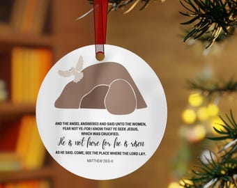 May include: Circular Christmas decoration with a religious theme. The ornament depicts a dove, a tomb, and text from Matthew 28:5-6, including "He is not here for he is risen." It is suspended by a red ribbon.