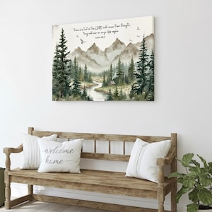May include: A framed watercolor print depicts a mountain landscape with a river, trees, and text from Isaiah 40:31. The print hangs above a wooden bench with pillows that read "welcome home". Green boots and a potted plant complete the scene.