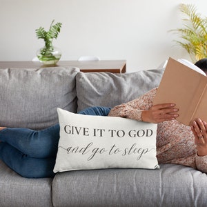 Puede incluir: Una almohada blanca con el texto "Give it to God and go to sleep" en letras negras. La almohada está en un sofá gris con una persona tumbada en él leyendo un libro.