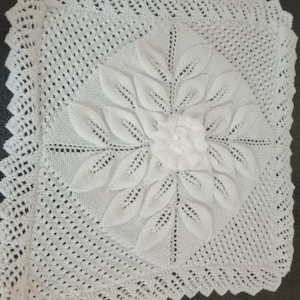 May include: A square, white, crocheted pillow cover featuring a central floral design. The design includes a large flower with detailed leaves and a textured center. The edges have a scalloped trim.