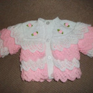 May include: A pink and white knitted baby cardigan with a scalloped edge and embroidered flowers.