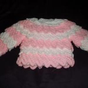 May include: A handmade baby sweater in a pink and white scalloped design. The sweater features long sleeves and a round neckline. The soft, textured yarn creates a cozy and visually appealing garment. The sweater is designed for infants.
