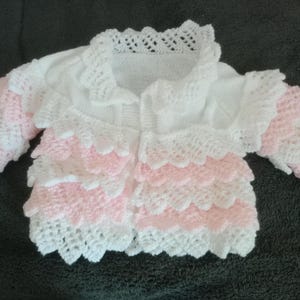 May include: A white and pink knitted baby cardigan. The cardigan features a scalloped design with white and pink layers on the sleeves and body. The collar is also white with a delicate lace pattern.