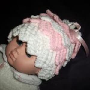 Può includere: Un berretto per neonato in crochet rosa e bianco con un fiocco bianco. Il berretto ha un design testurizzato e arricciato.