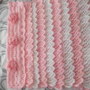 May include: A handmade, pink and white crochet blanket with a scalloped edge. The blanket features a repeating pattern of pink and white stripes. A section of the blanket has three pink, crocheted flowers.