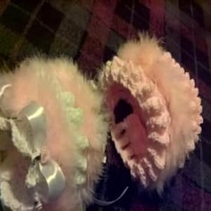 May include: A pair of baby booties in a soft pink colour, decorated with fluffy feathers and delicate white bows. The booties have a crocheted edge and are designed for infants. The background is a patterned fabric.