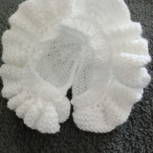 Puede incluir: Un gorro blanco tejido a mano con un borde con volantes. El gorro está hecho de hilo suave y tiene una forma circular con una abertura para la cara. El borde con volantes añade un toque decorativo al accesorio para bebés.