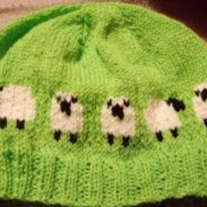 May include: A bright green knitted beanie featuring a row of white sheep with black faces and legs. The hat is made of a textured yarn and has a rounded crown. The sheep are evenly spaced around the lower edge of the hat.