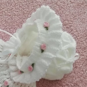 May include: White knitted baby bonnet with a ruffled edge and pink flower embellishments.