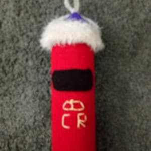 Puede incluir: Un buzón de juguete de punto rojo con ribete de piel blanca y parte superior azul. El buzón tiene una abertura negra y la inscripción "E R" en la parte delantera.