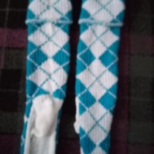 Puede incluir: Un par de calcetines hasta la rodilla con un patrón de rombos blanco y turquesa. Los calcetines tienen un puño doblado en la parte superior y están hechos de un material de punto suave. El patrón de rombos es un diseño clásico.