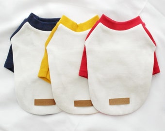 May include: Three dog shirts in white with colored trim. The shirts have a navy blue, yellow, and red trim. Each shirt has a small brown label with the text "Honey Dog" on it.