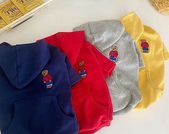 May include: Four hooded dog sweaters in different colors: navy blue, red, gray, and yellow. Each sweater features a small embroidered bear with a blue shirt and red pants.