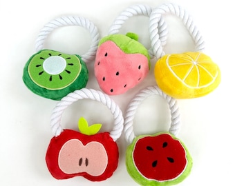 Può includere: Cinque giocattoli di peluche a forma di frutta colorati con manici in corda bianca. I frutti sono un kiwi, una fragola, un limone, una mela e un'anguria.