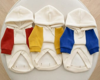 May include: Three white hooded dog sweaters with different colored sleeves. The sleeves are red, yellow, and blue.
