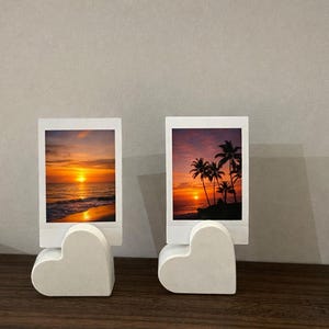 May include: Two white heart-shaped photo holders displaying sunset images. Each holder has a white frame holding a photo of a sunset over the sea. The sunset features orange, yellow, and red colours.